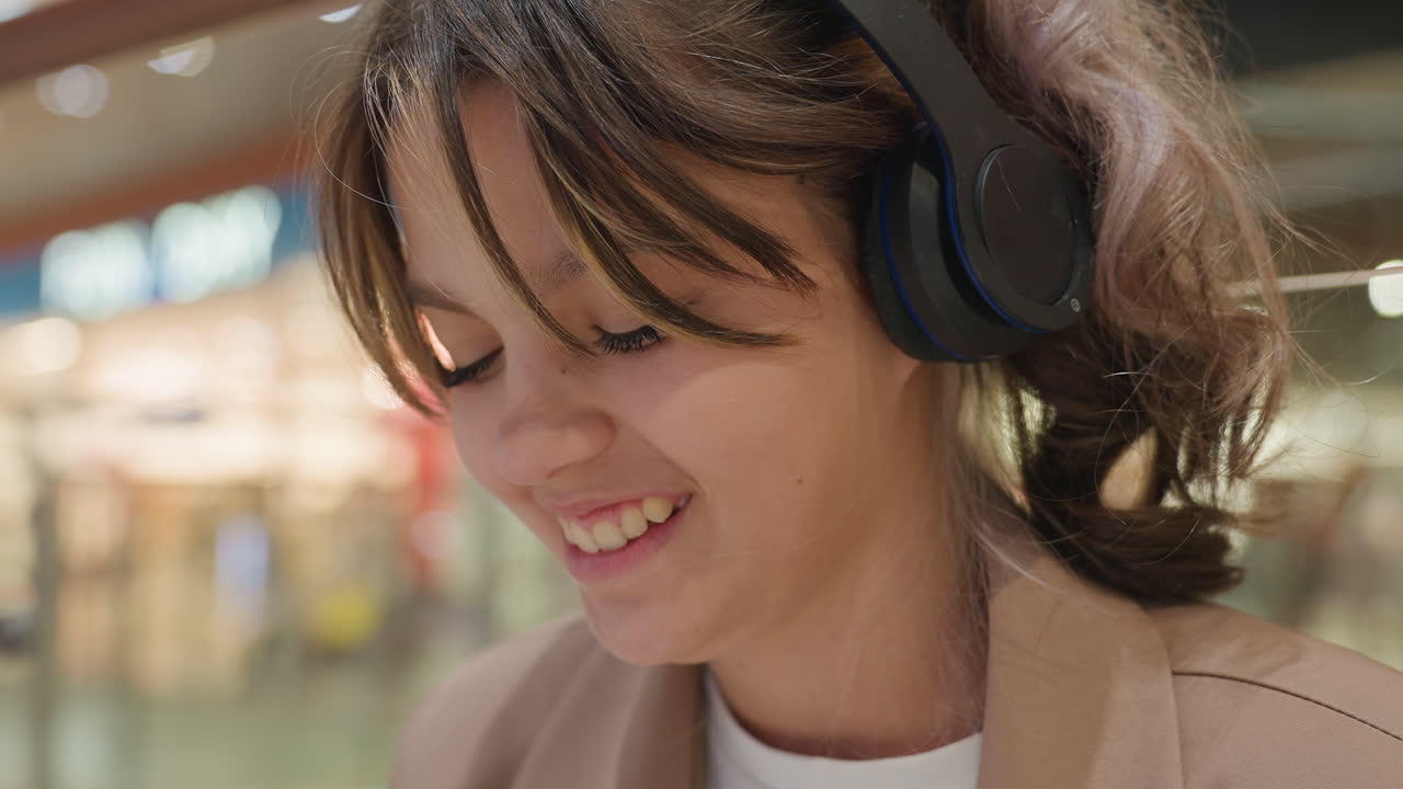 음악 쇼핑을 즐기는 젊은 여성, 아늑한 조명 아래에서 편안하게 미소 지으며 음악 재생 목록을 둘러보는 소녀, 번화한 쇼핑 센터에서 베이지색 재킷을 입고 편안하게 음악을 즐기는 즐거운 젊은 여성