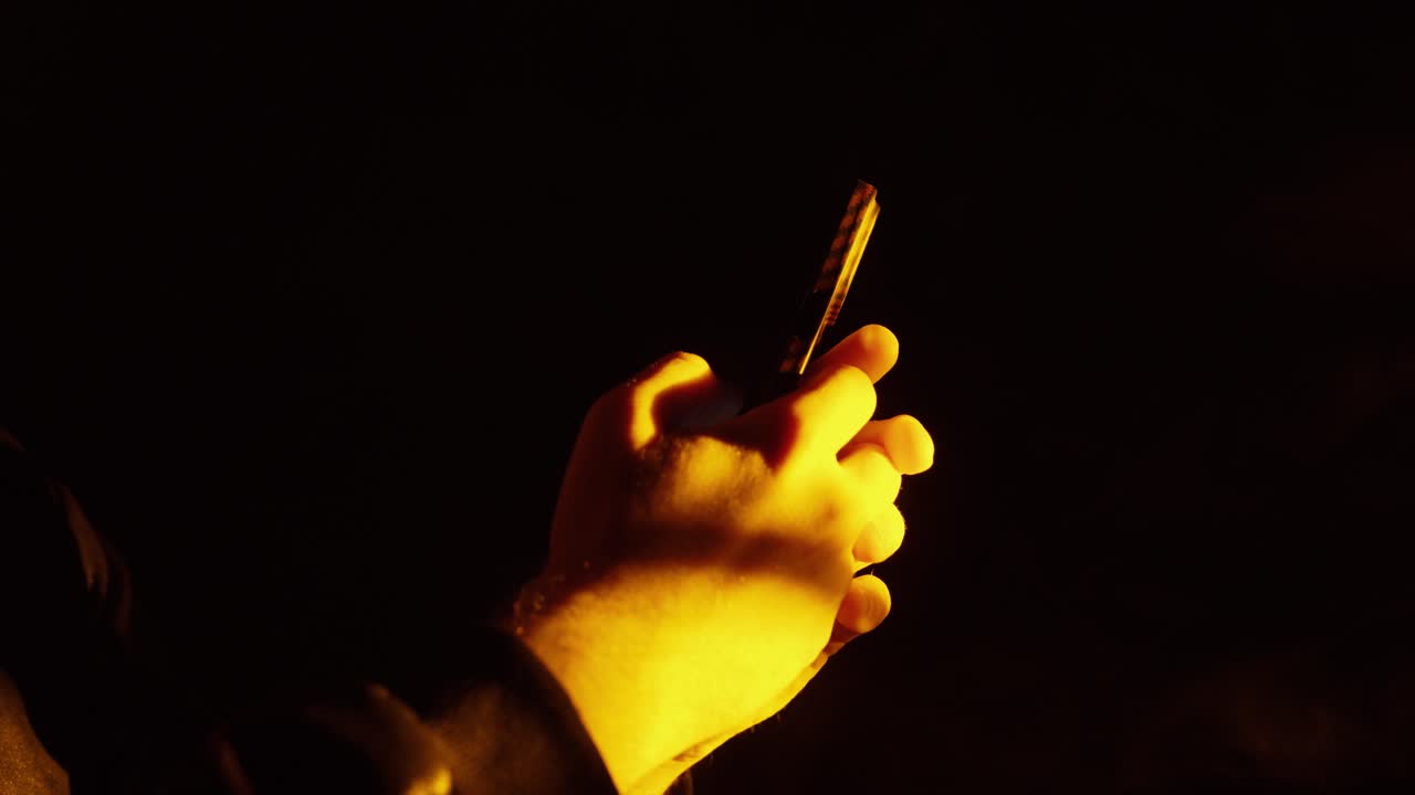 Cinematic close-up of hands holding a smartphone at night in warm orange light, creating a moody atmosphere and symbolizing modern digital lifestyle