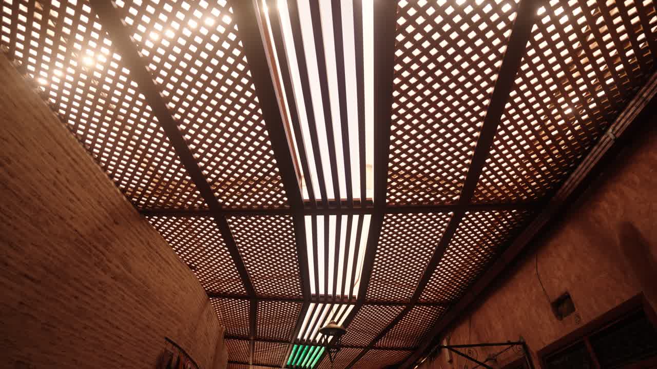 A wooden lattice roof with hanging lanterns filters sunlight inside a traditional Moroccan market