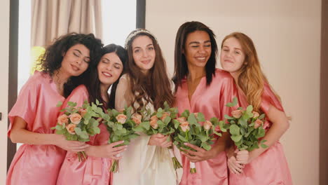 group of multiethnic female friends and bride looking at camera, wearing silk pink and white nighdresses while holding bouquets 1