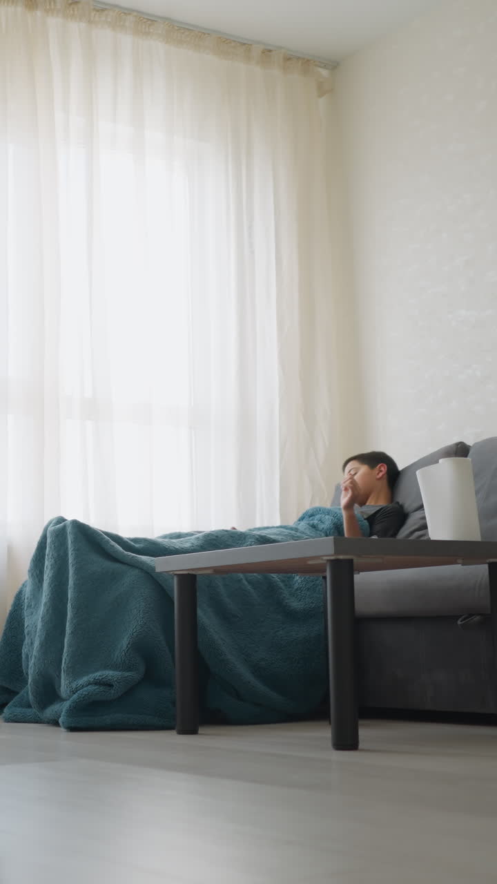 Boy covered in blanket coughs while lying on sofa in bright modern room, covering mouth with hand, resting quietly beside tissue box on table, suggesting cold symptoms