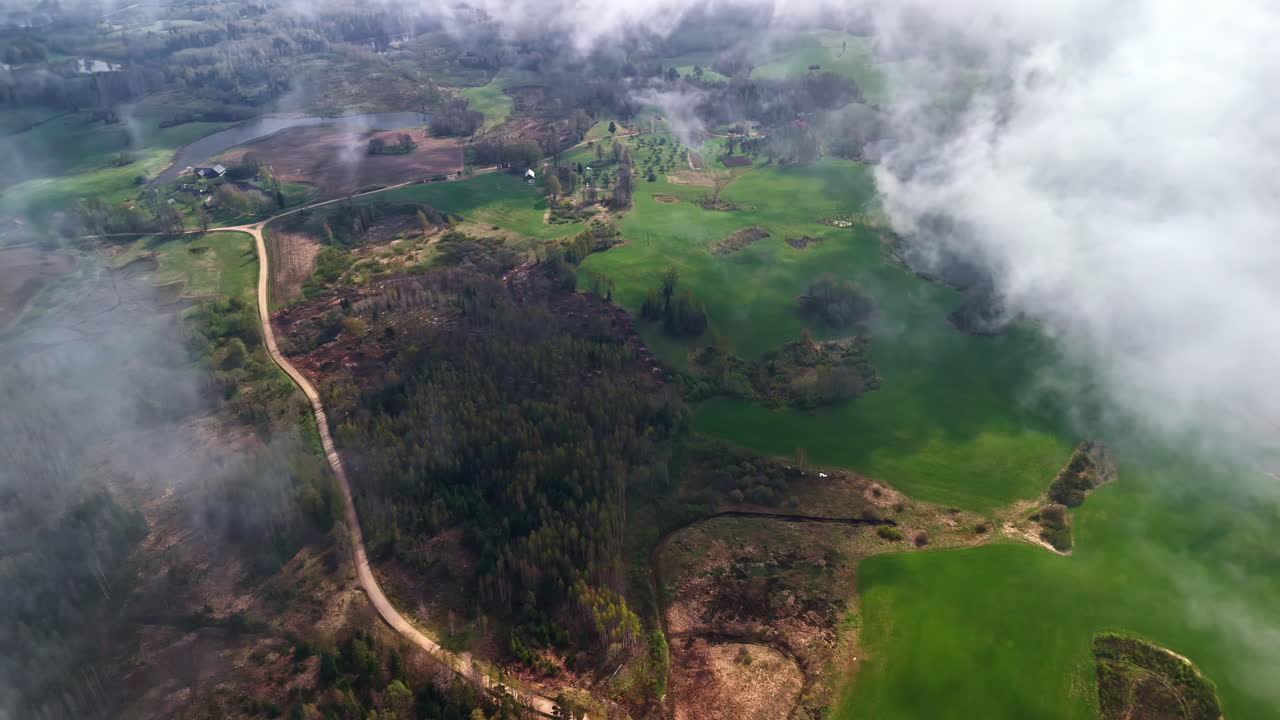 Aerial flight through white clouds looking down on winding country road through lush green fields and forests in Latvia