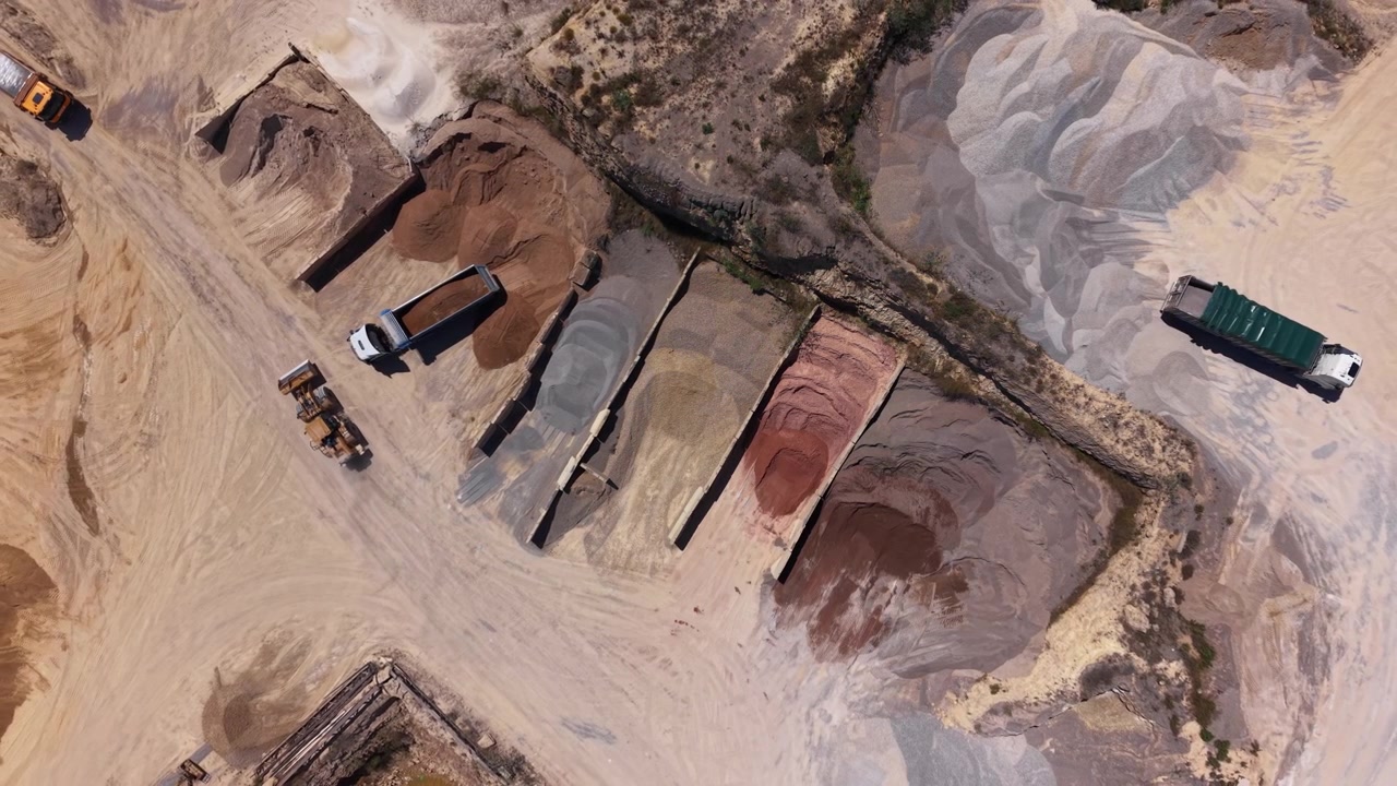 Overhead view of an active sand and gravel quarry with dump trucks, a loader, and colorful mineral stockpiles arranged across a dry excavated landscape