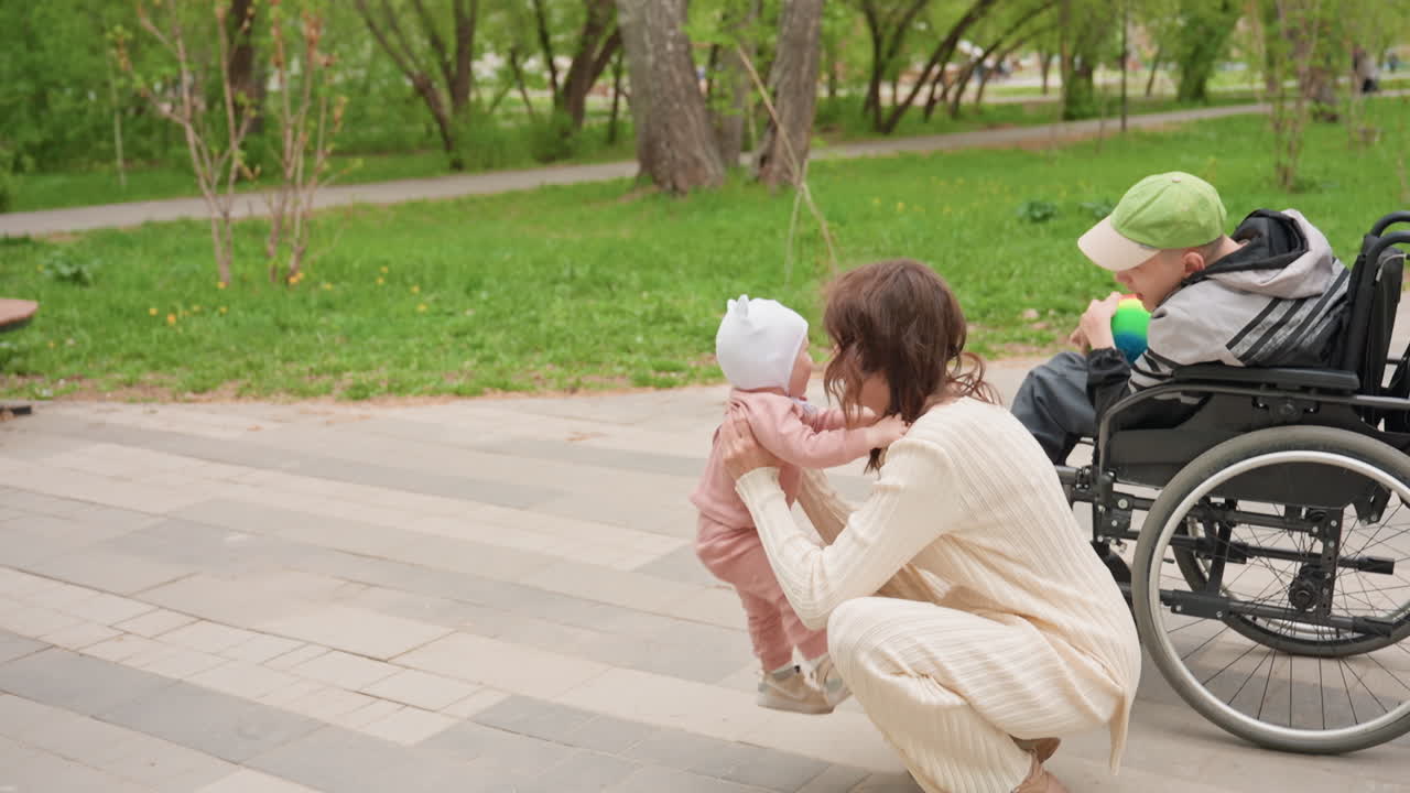 Paved Plaza Toddler Taking First Steps Toward Reaching Hands, Parent Crouched To Encourage, Wheelchair And Companion Nearby, Joyful Milestone, Urban Park Setting, Supportive Cheer, Bright Spring Day