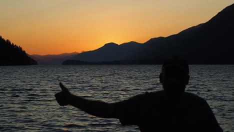 Puesta-De-Sol-En-Un-Lago-Con-Montañas-En-El-Fondo-Y-Un-Hombre-Caminando-Dando-Los-Pulgares-Hacia-Arriba