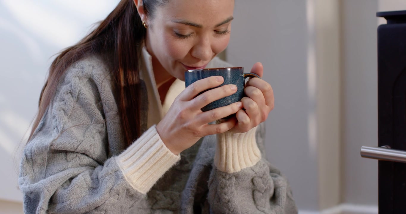 Inquadratura allargata che rivela una donna asiatica in maglione seduta a casa con regali, che tiene una tazza e sorseggia per scaldarsi.