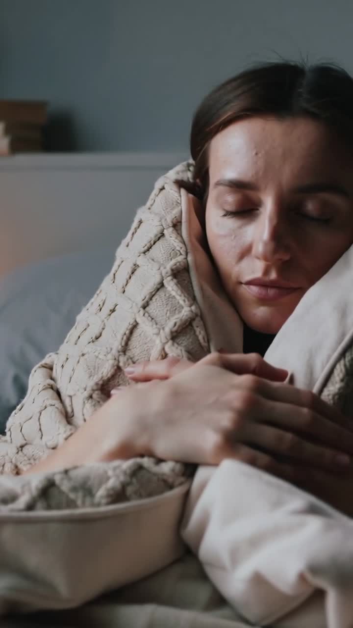 Video verticale: Donna che allenta la presa, si rilassa sul letto, tenendo una coperta color crema e un cuscino per riposare.