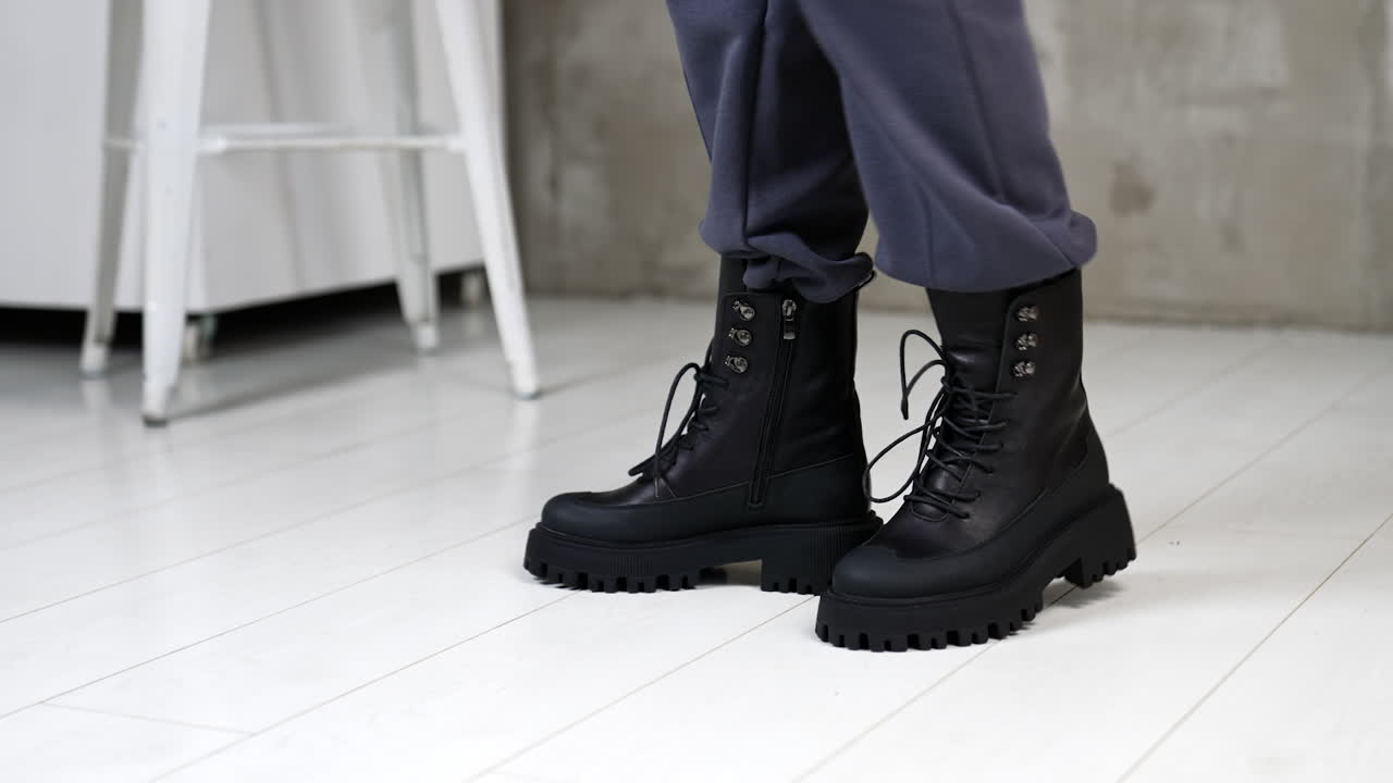 Demonstrating the black stylish leather boots with tractor soles. Female in grey loose pants presenting the footwear with laces for autumn season.