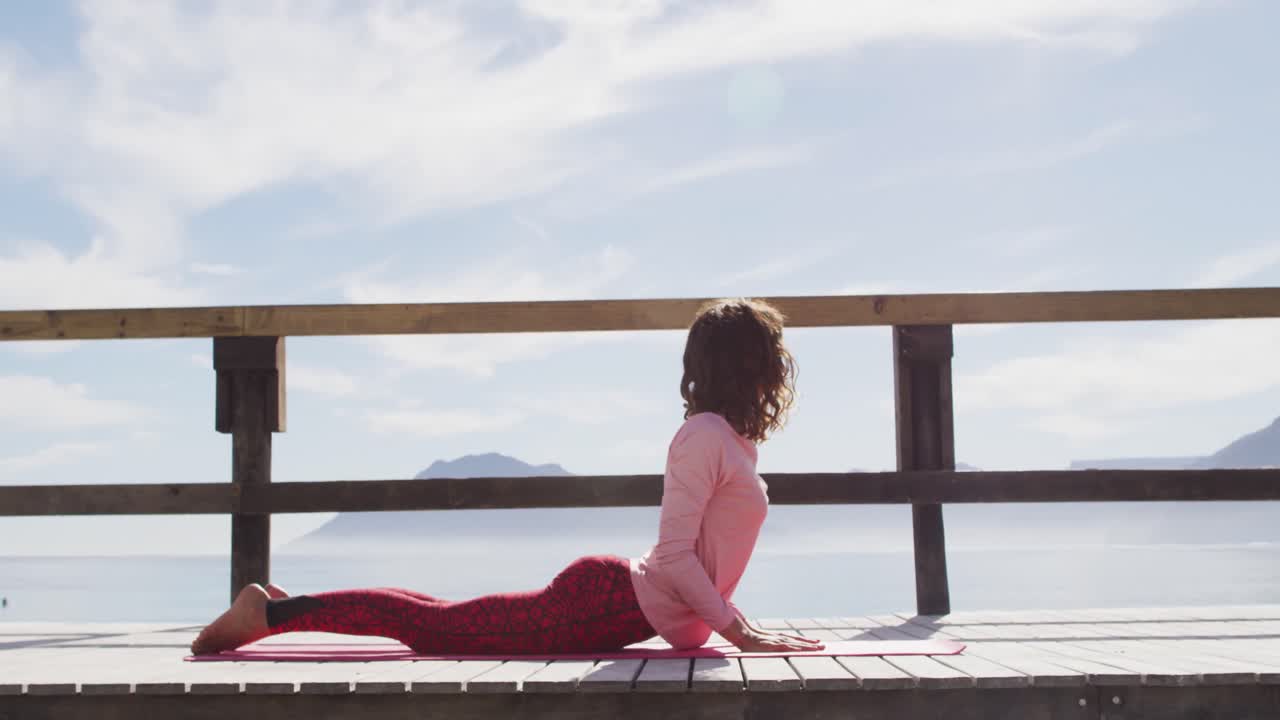건강 한 인종 의 여성 이 야외 에서 요가 를 연습 하고, 바 ⁇ 가 에 누워서 스트레칭 하고 있다
