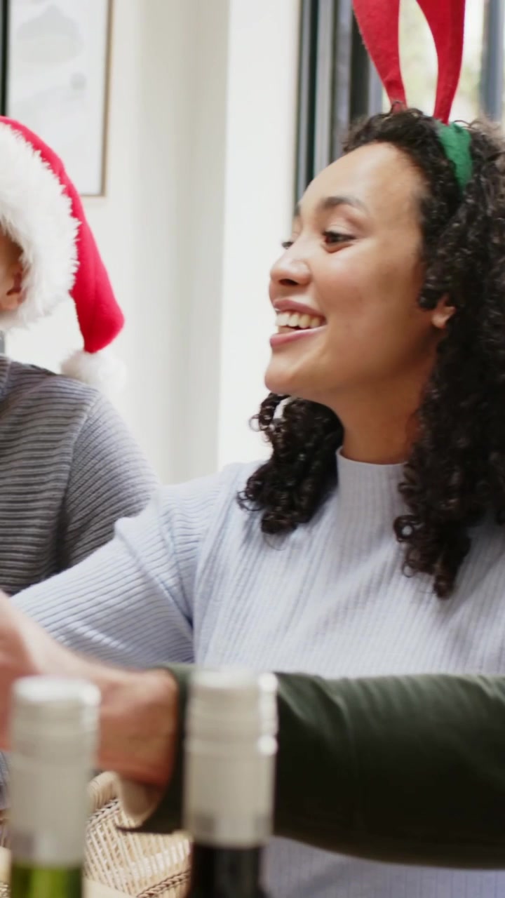 Vídeo vertical: Tilintar de copos enquanto mulher com tiara de chifres levanta o copo à mesa com homem de gorro de Papai Noel brindando ao feriado.