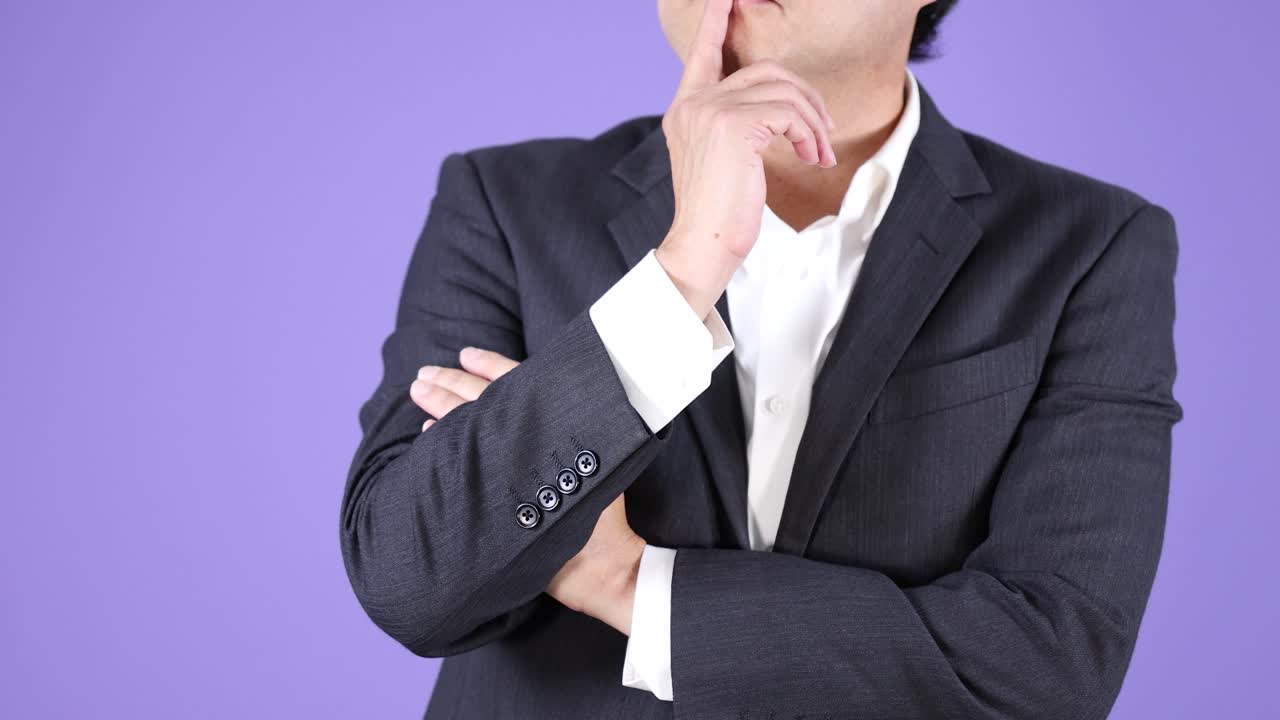 スーツを着たプロフェッショナルな男性が、均一な照明の下で、顎に手を当てて熟考している、静的なミディアムショット