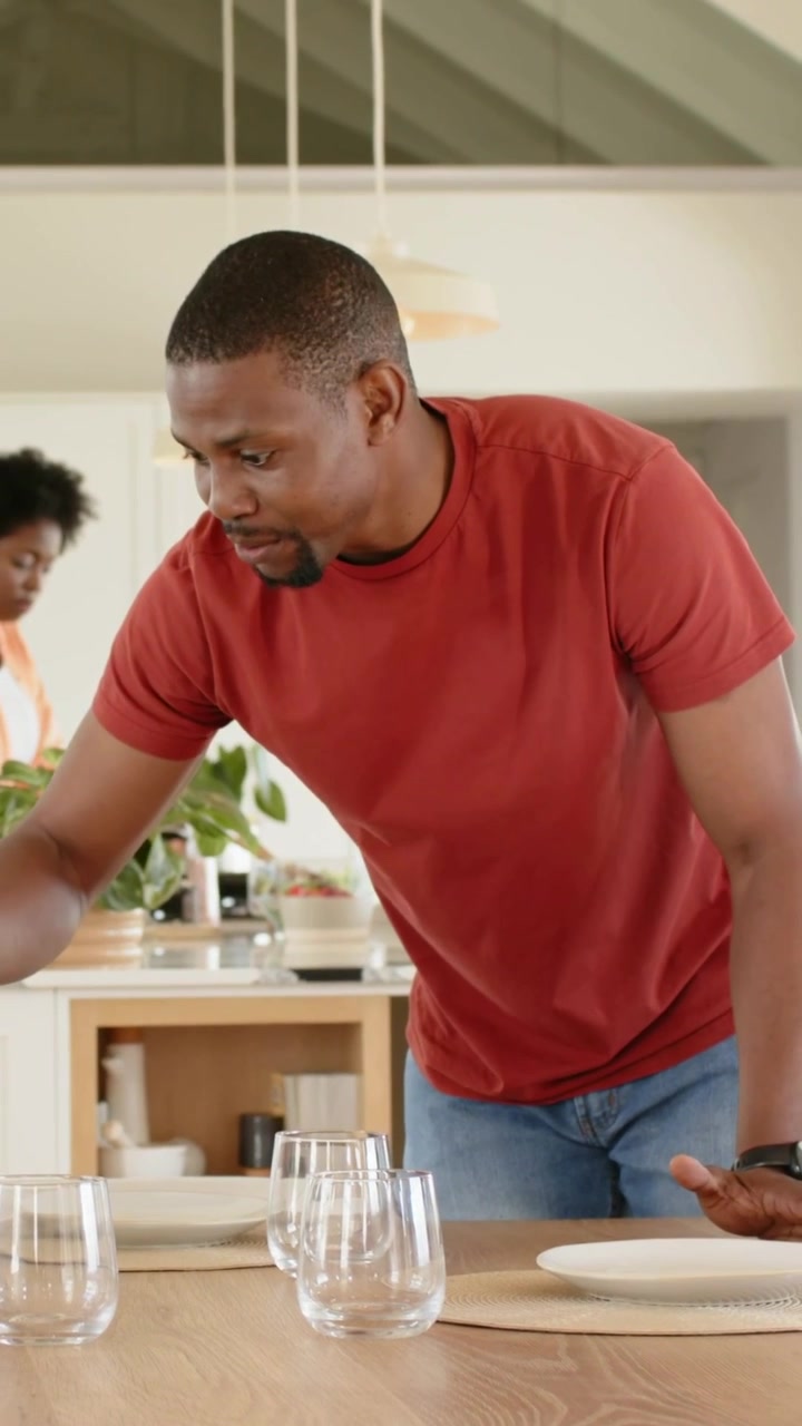 Vertical video: Leaning, man wearing red tee placing plates, glasses on home table, preparing meal