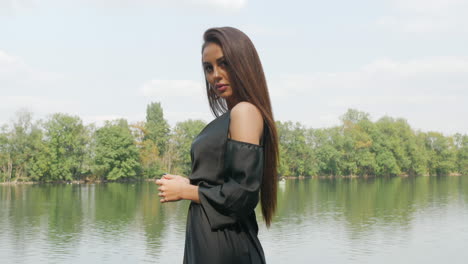 young russian lady wearing a black dress, stands in front of a lake, surrounded by trees