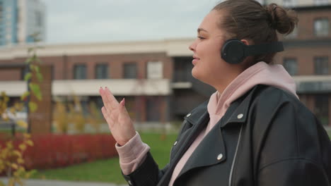 Joyful lady dances in outdoor urban setting enjoying music through wireless headphones while autumn breeze flows gently around buildings and colorful foliage sways in background