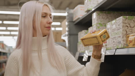 housewife with platinum hair strolls grocery aisle, lifts boxed vegan soap from shelf, studies price label, weighing household budget, warm lights reflecting off stacked cartons