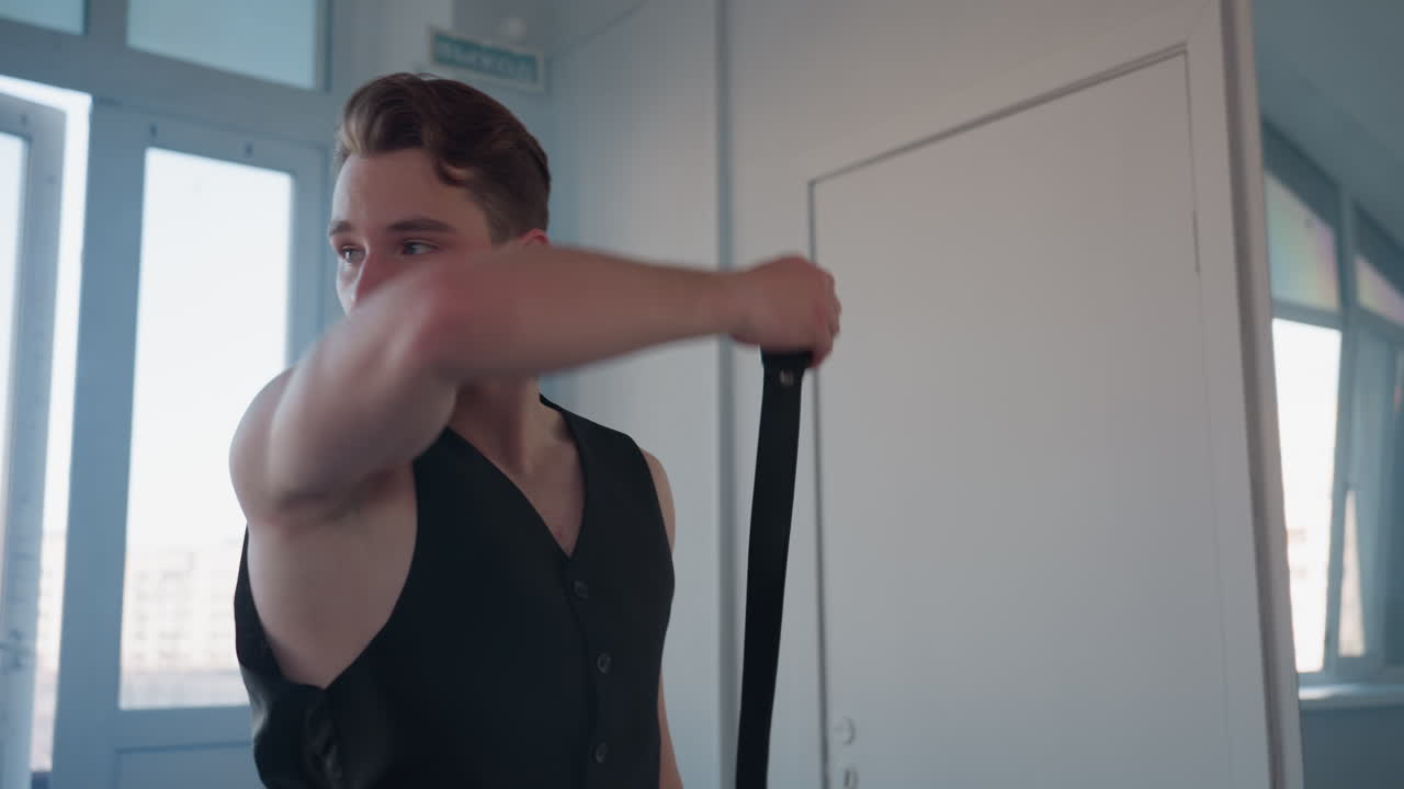 white young man drapes belt around neck while blowing kiss in confident dance move inside sunlit loft studio framed by tall windows mirror reflection