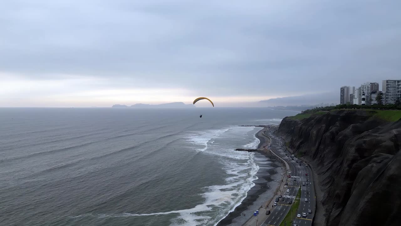 Imagem aérea orbital mostrando um parapentista fazendo curvas para perder altitude e se aproximar do pouso, com uma ilha e, em seguida, edifícios da cidade ao fundo.