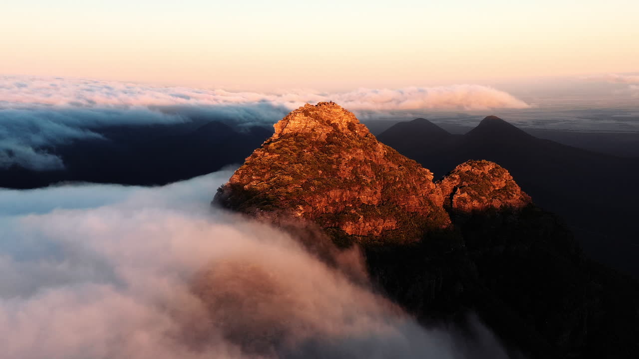 Aerial, drone shot over clouds towards a sunlit summit of a rocky mountain, during sunset, in Hassell National Park, West Australia