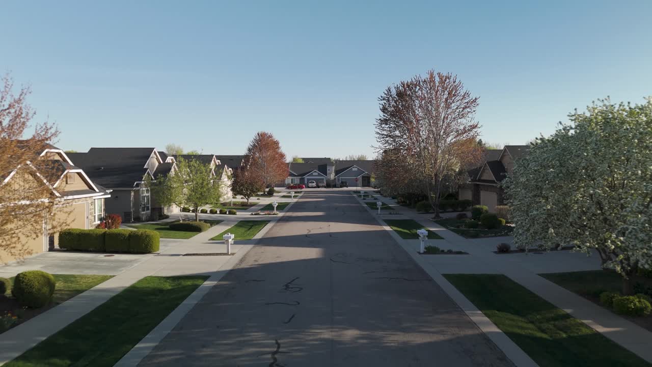 Drone aerial of a quiet suburban street, in Spring, lined with well-kept homes, manicured lawns, and blossoming trees, clear blue sky, in a residential neighborhood