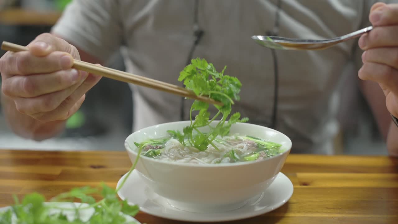 Bát phở bò Việt Nam bốc khói trên bàn gỗ, du khách da trắng nếm thử nước dùng bò trong veo bằng thìa và đũa, rau mùi tươi cùng các loại rau thơm khác được đặt bên cạnh, trong không khí quán ăn vỉa hè bình dị.