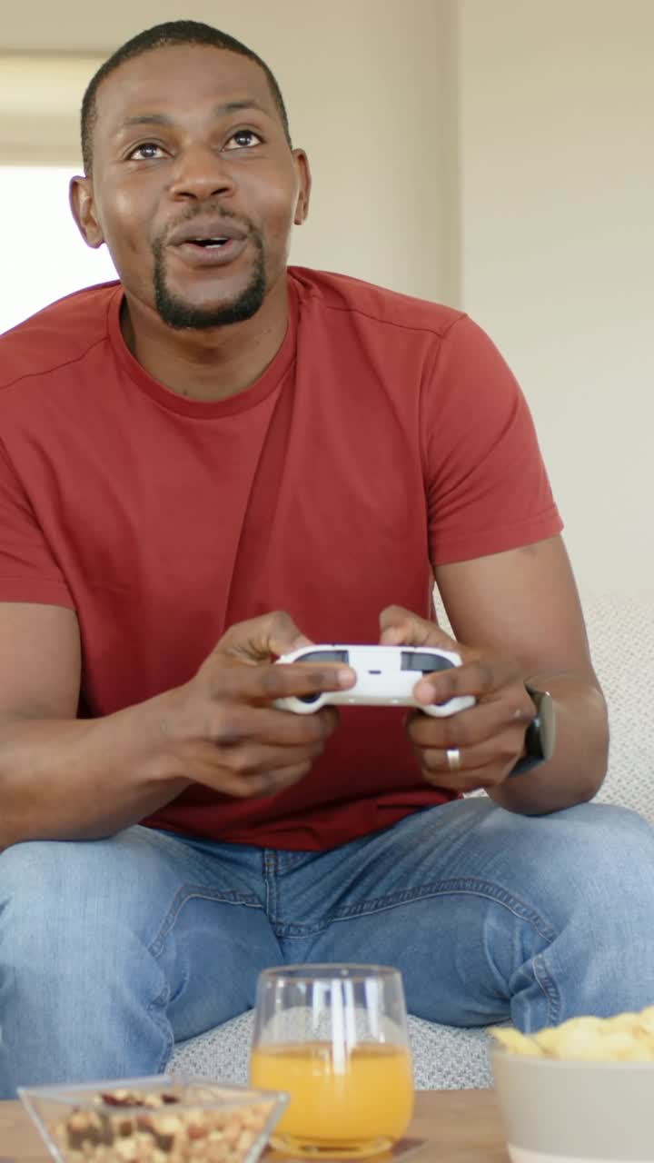 Vertical video: Smiling then playing, man in red tee and jeans operating white controller on couch