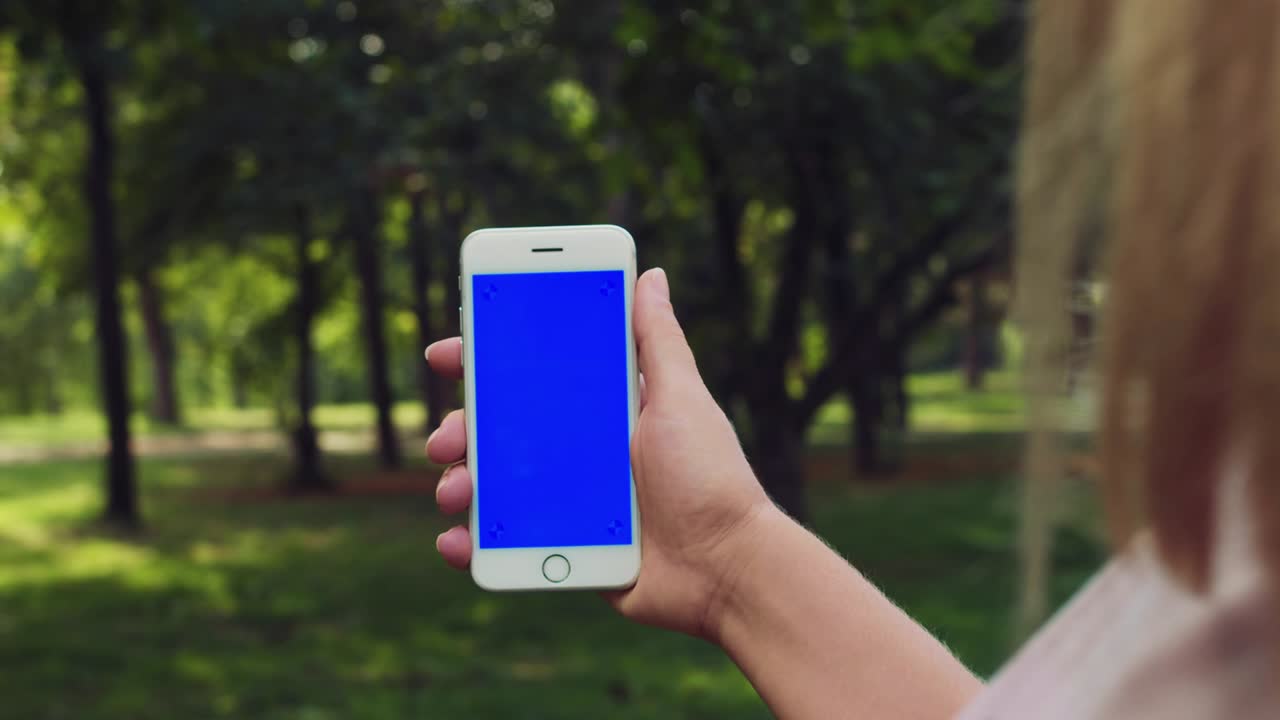 cerca de la mano de una mujer joven que sostiene un teléfono inteligente con pantalla azul tecla de croma en el fondo de hierba verde al aire libre texto celular de teléfono celular dedo en cámara lenta