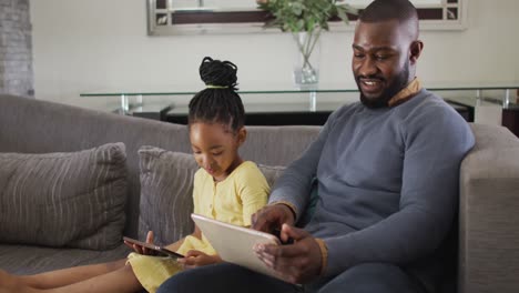 happy african american daughter and father using tablets on sofa