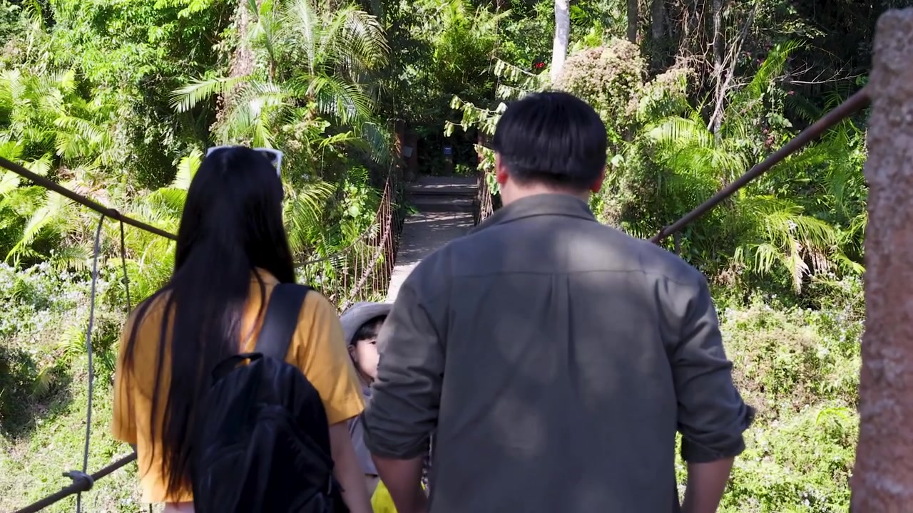 Seorang pria dan wanita berjalan menjauh dari kamera melintasi jembatan gantung hutan