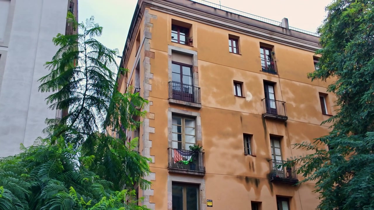 Edificio residenziale tradizionale a Barcellona, Catalogna, Spagna, con pareti in stucco color ocra, finestre alte e balconi in ferro battuto incorniciati da alberi urbani maturi, panoramica della telecamera.