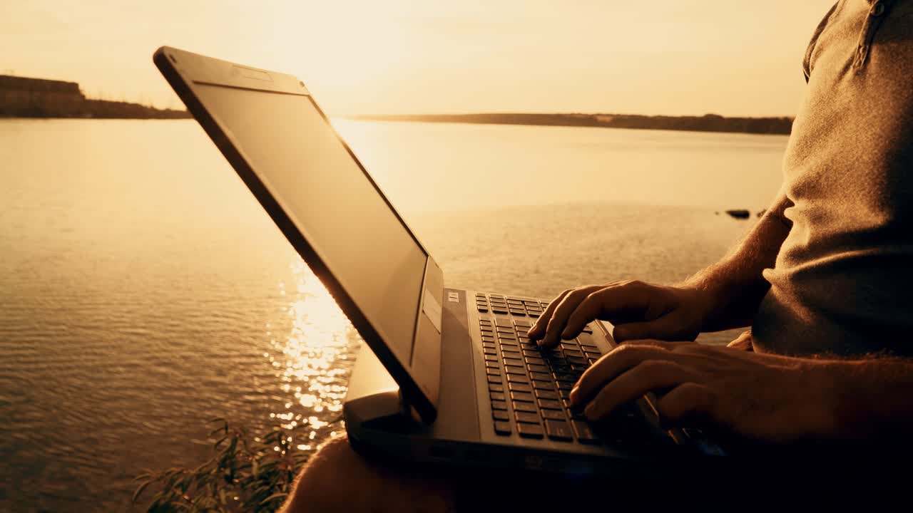 Man's hands using laptop computer on top of a hill by the river. Beautiful summer sunset