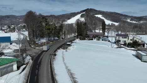 push in drone shot flying above snow covered town, truck driving on the road below, with ski area and mountain town in distance