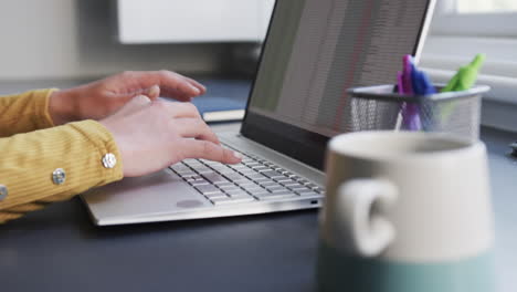 sección media de una mujer biracial sentada en un escritorio usando una computadora portátil y bebiendo café en casa, en cámara lenta