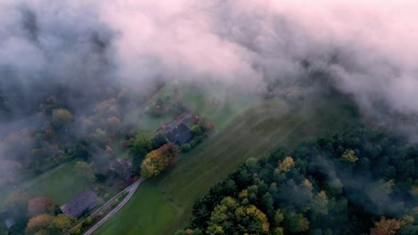 an aerial view of a dense, green forest with patches of sunlight breaking through the fog, revealing several houses with brown roofs nestled among the trees