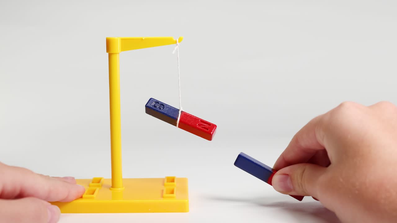 Hands manipulate magnets to demonstrate attraction and repulsion using a suspended bar magnet on a yellow stand