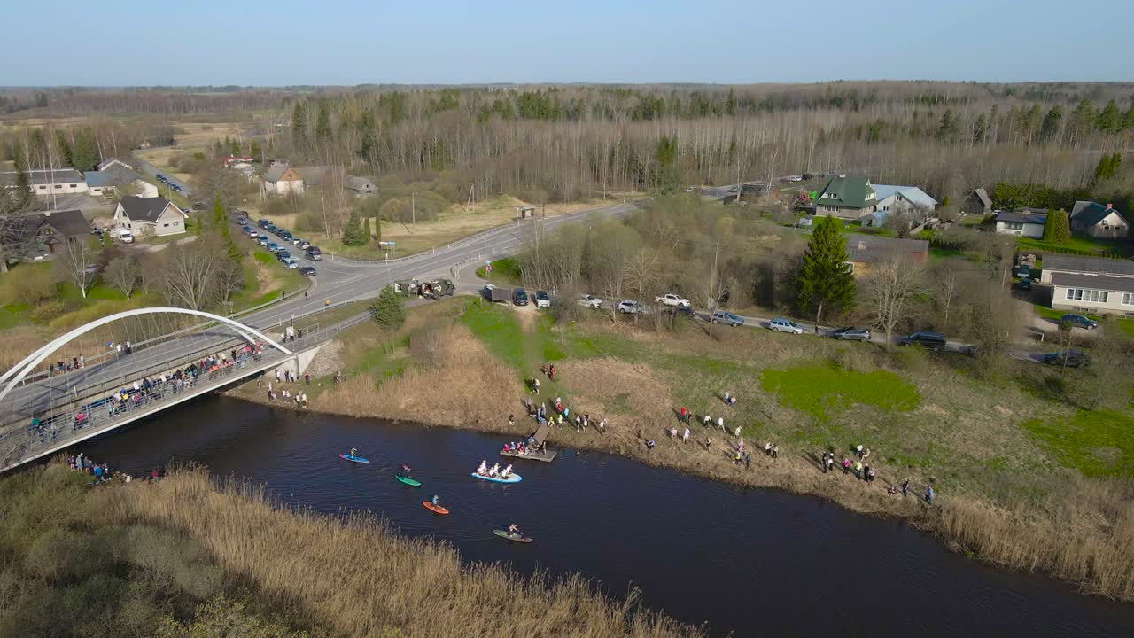 Aerial drone footage of kayakers, SUP paddleboars and rafts paddling along a river from under a bridge during Võhandu marathon while spectators watch them on grassy and sunny green shorelines.