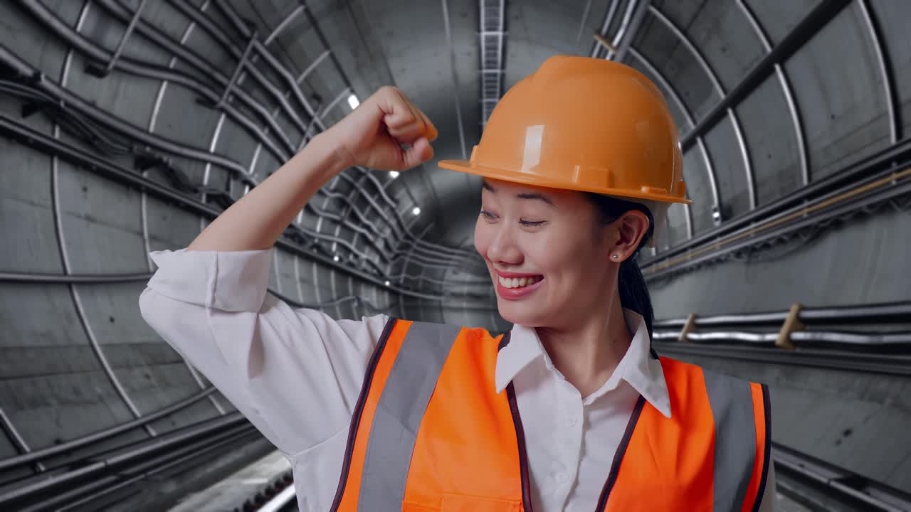 Close Up Of Asian Female Engineer With Safety Helmet Flexing Her Bicep And Smiling To Camera In Underground Subway Tunnel