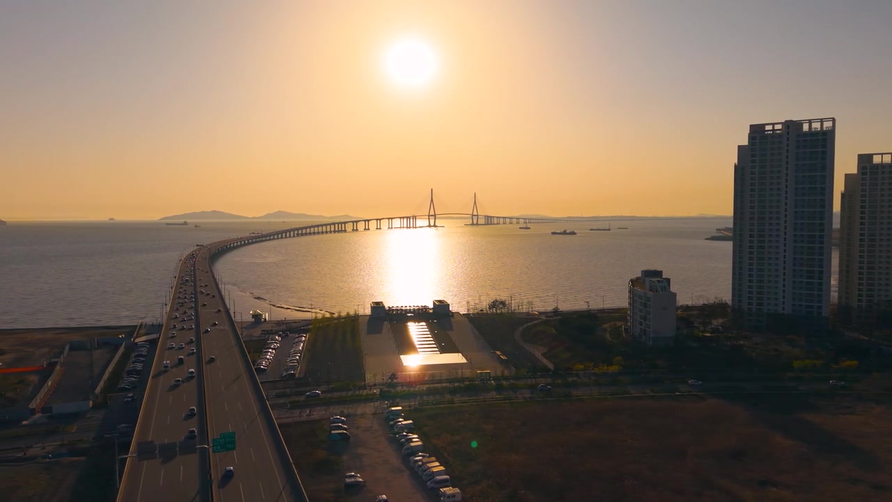 Un volo aereo laterale lungo il sinuoso Ponte di Incheon cattura il bagliore dorato del tramonto sull'oceano e sulle torri di appartamenti di Songdo.