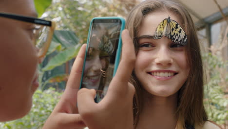 Wunderschöner-Schmetterling-Auf-Dem-Gesicht-Eines-Mädchens-Mit-Einem-Glücklichen-Freund,-Der-Mit-Dem-Smartphone-Fotografiert.-Freunde-Haben-Spaß-Im-Zoo-Wildreservat-Und-Teilen-Naturausflüge-In-Sozialen-Medien-Mit-4K-Videos