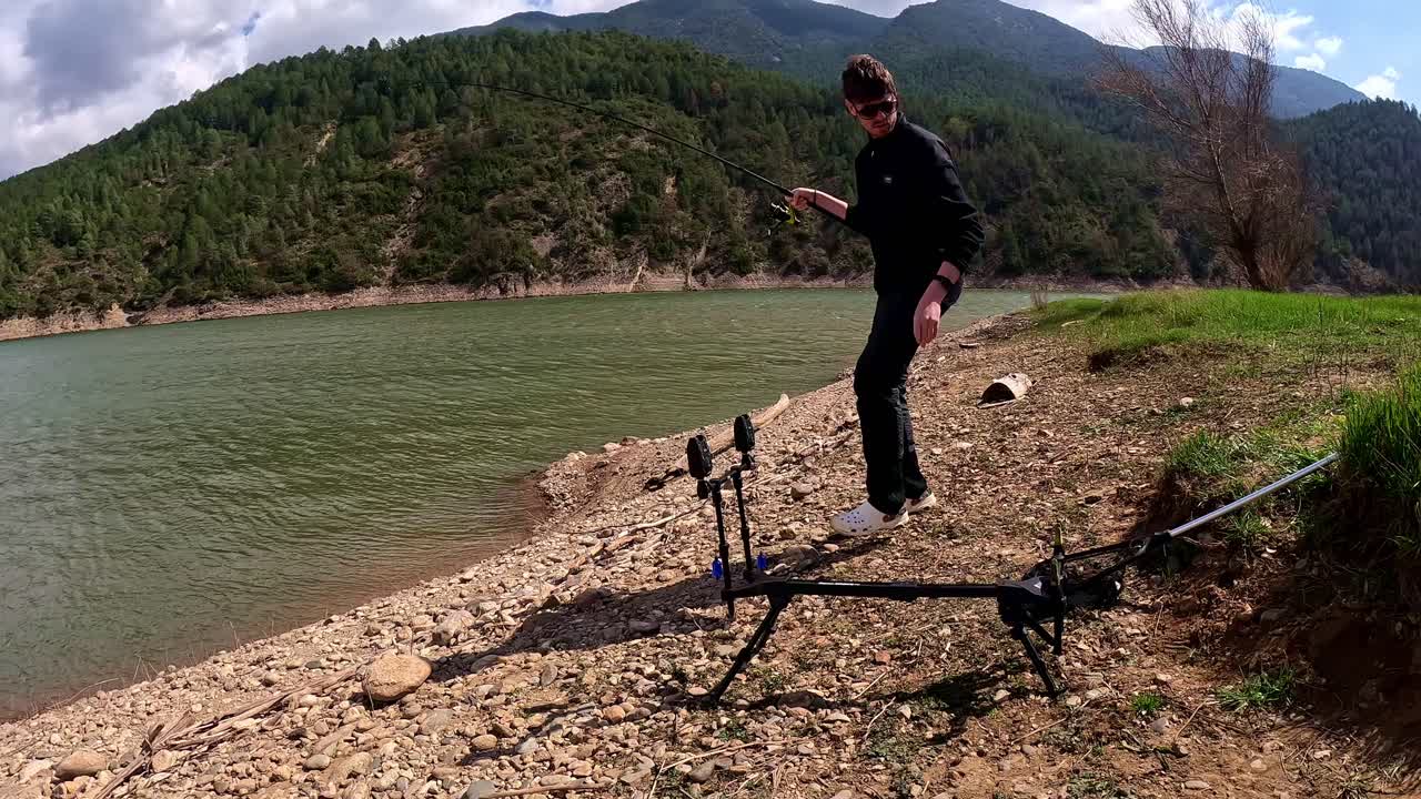 A fisherman is poised on a rocky lakeshore, using a tripod for carp fishing. He is ready with a landing net to retrieve a fish from the water. The backdrop features a scenic view of forested hills