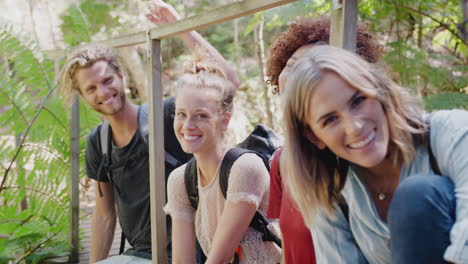 Portrait-Of-Young-Friends-Hiking-Through-Countryside-Sitting-On-Wooden-Bridge-Together