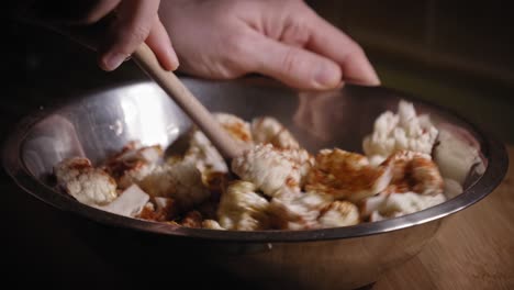 person are using wooden spoon to mix cauliflower with salt, spices and oil - handheld shot