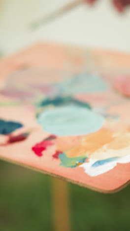 un artista con una camisa a cuadros, con su cara no visible, sostiene un pincel y una paleta llena de colores vibrantes. el fondo borroso revela un lienzo blanco