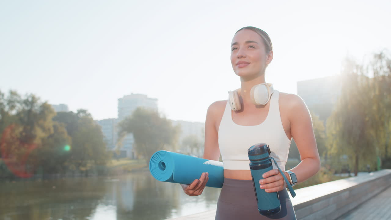 Happy young sportswoman jogger with water bottle and rolled mat walking in city park on sunny day