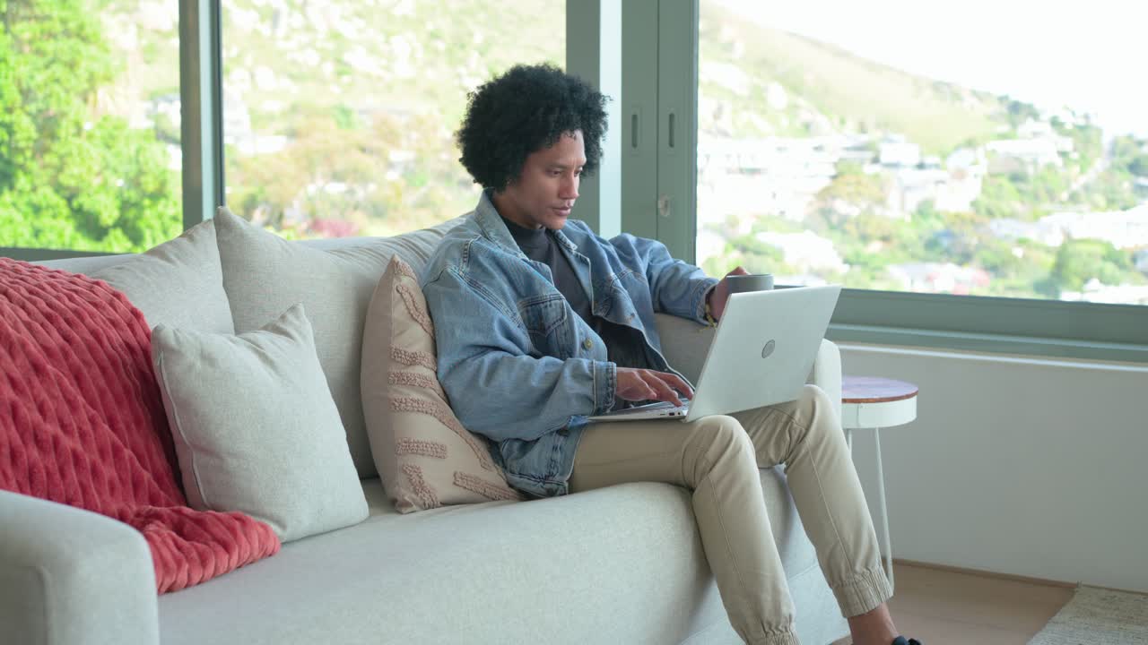 Man sipping coffee mug on sofa in living room, working on laptop, glancing at hillside