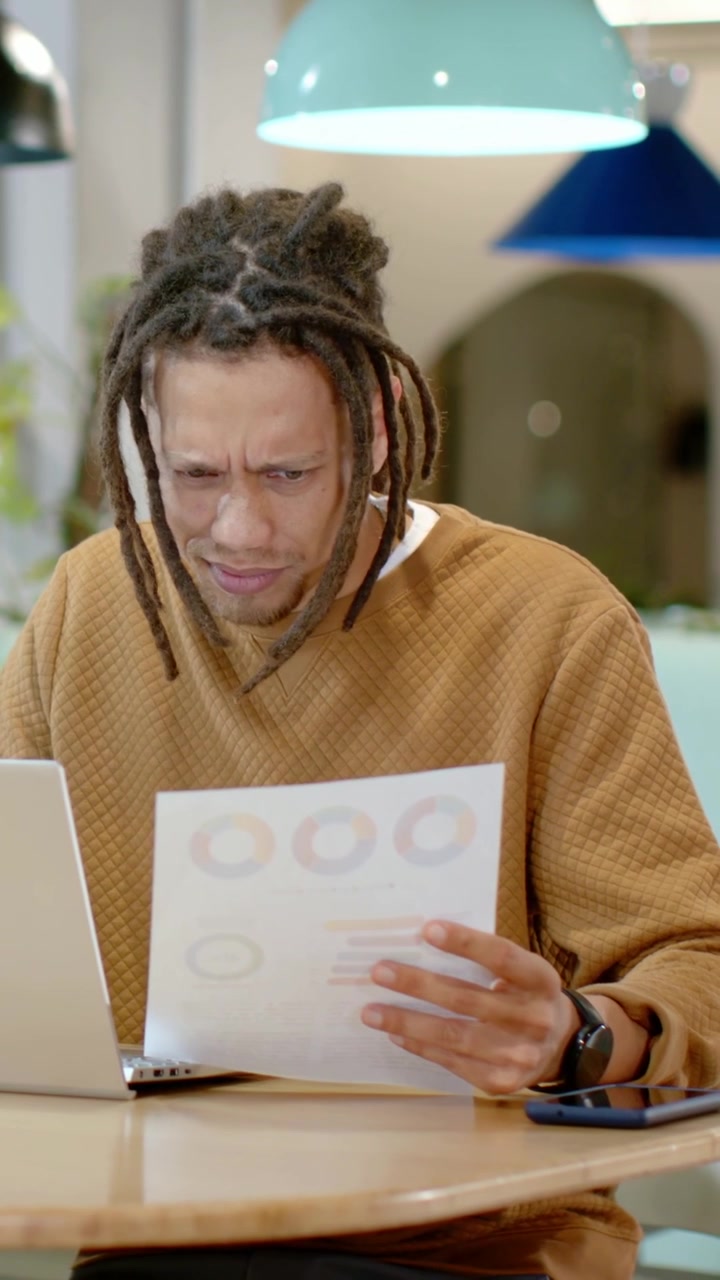 Man lifting printed charts and comparing to laptop screen for business analysis in coworking lounge
