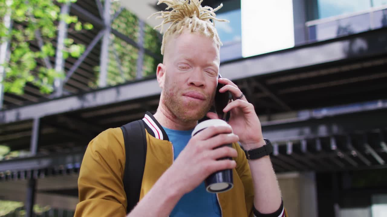 feliz albino hombre afroamericano con rastas bebiendo café hablando en el teléfono inteligente