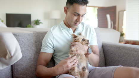 happy biracial man sitting on sofa, petting pet yorkshire terrier dog and smiling, slow motion