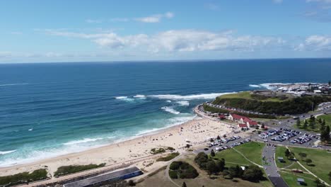 Drone-flying-towards-the-ocean-over-a-beach,-people-swimming-below