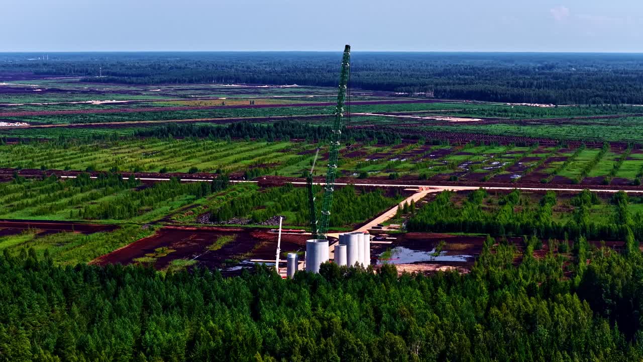 Crane constructs wind turbines in front of vast green fields and dense forest landscape