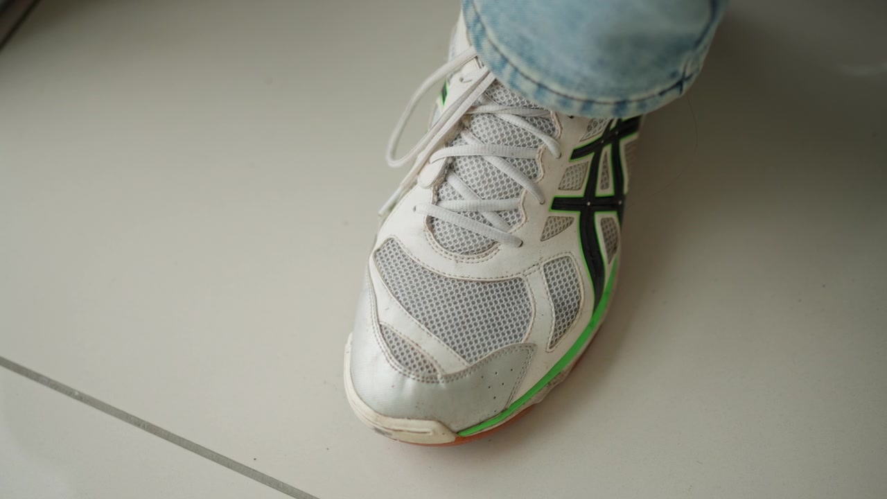 Primo piano di una sneaker bianca con dettagli verdi indossata su un piede, che mostra la texture dettagliata in mesh e i lacci su un pavimento di colore chiaro. Il piede rimane fermo, enfatizzando gli elementi di design della scarpa.